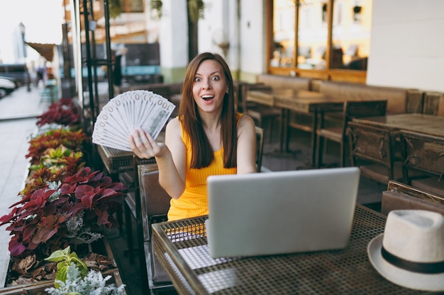 Femme au café de la rue en plein air assis avec un ordinateur portable moderne, tient dans la main un tas de billets de banque en dollars