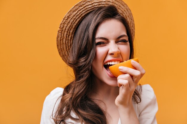 Femme au chapeau de paille et haut blanc mord l'orange avec les yeux fermés.