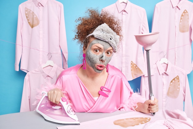 Une femme au foyer bouclée surprise subit des soins de beauté tout en faisant le ménage applique un masque d'argile sur le visage tient des fers à repasser sur des vêtements ou du linge sur une planche à repasser porte un masque de sommeil et une robe de chambre