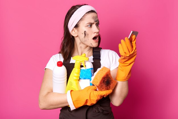 Femme au foyer brune élancée choquée détient des bouteilles de détergent de nettoyage dans une main et un téléphone dans l'autre