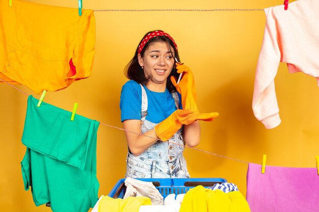 Femme au foyer drôle et belle faire des travaux ménagers isolés sur l'espace jaune. Jeune femme caucasienne entourée de vêtements lavés