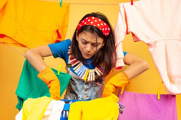 Femme au foyer drôle et belle faire des travaux ménagers isolés sur fond jaune. Jeune femme caucasienne entourée de vêtements lavés. Vie domestique, œuvres d'art lumineuses, concept d'entretien ménager. Ça a l'air occupé.