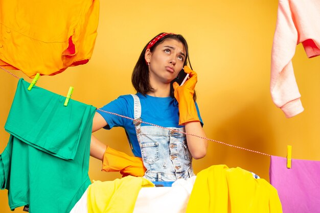 Femme au foyer drôle et belle faire des travaux ménagers isolés sur fond jaune. Jeune femme caucasienne entourée de vêtements lavés. Vie domestique, œuvres d'art lumineuses, concept d'entretien ménager. Parler au téléphone.