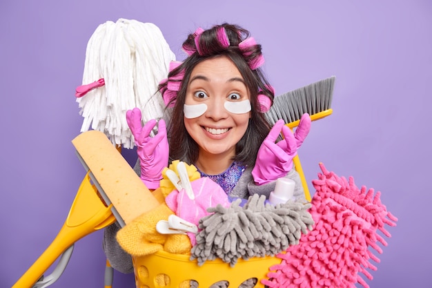 Une femme au foyer ethnique heureuse lève les mains en souriant utilise largement des produits de nettoyage et le matériel d'entretien ménager applique des tampons sous les yeux rend la coiffure isolée sur fond violet. Concept de ménage