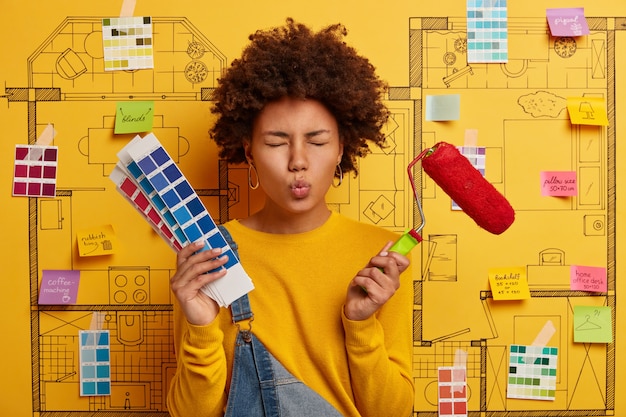 La femme au foyer garde les lèvres arrondies, s'occupe de la rénovation de la maison, tient le pinceau et la palette de couleurs, effectue la réparation dans un appartement en fonction du projet de conception. Peintre en bâtiment pose sur croquis sur mur jaune