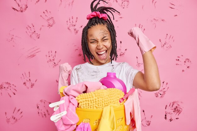 Une femme au foyer heureuse fait un geste oui serre les poings célèbre la fin du nettoyage de la maison porte des gants en caoutchouc se tient près du panier à linge s'amuse isolé sur un mur rose sale