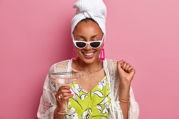 Femme au foyer joyeuse s'amuse à la soirée pyjama, lève le poing fermé, célèbre un événement spécial, boit un cocktail, porte une serviette de bain sur la tête, des lunettes de soleil, glousse positivement, isolé sur un mur rose