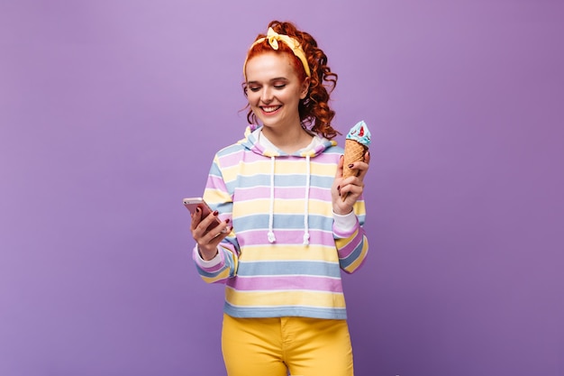 Une femme au gingembre en coiffure jaune avec un sourire discute dans un smartphone et tient une glace bleue