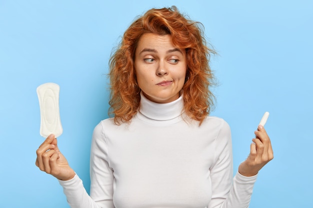 Une femme au gingembre hésitante regarde de manière confuse une serviette hygiénique et un tampon