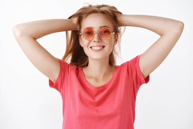 Femme au gingembre insouciante et mignonne avec des taches de rousseur et un large sourire mignon tenant les cheveux derrière avec les deux mains regardant satisfait et insouciant de détente