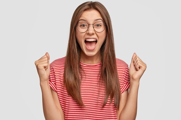 Femme autodéterminée aux cheveux raides foncés, serre les poings et s'exclame avec une expression de triomphe