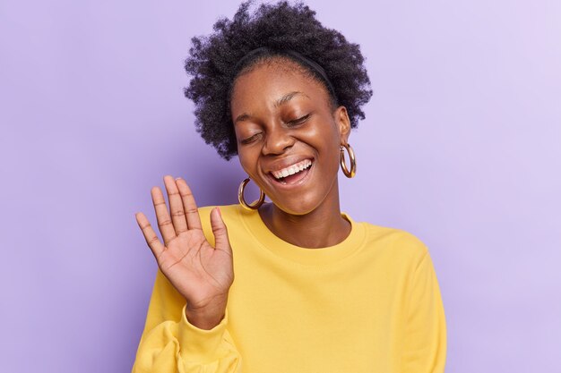 femme aux cheveux afro garde la paume levée ferme les yeux sourires montre positivement les dents isolées sur violet