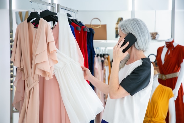 Femme aux cheveux blonds parlant au téléphone mobile tout en choisissant des vêtements et en parcourant des robes sur une grille dans un magasin de mode. Coup moyen. Client de boutique ou concept de vente au détail
