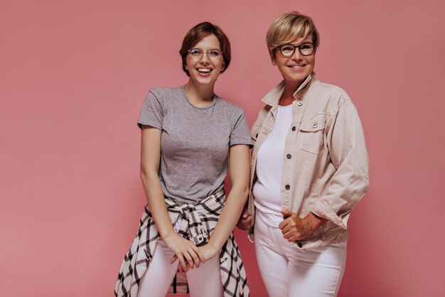 Femme aux cheveux blonds en veste beige et lunettes souriant et posant avec une jeune fille en t-shirt gris et chemise à carreaux sur fond rose.