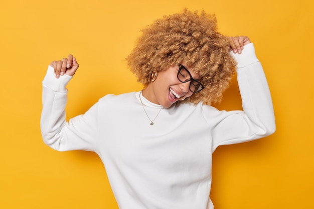 Photo gratuite une femme aux cheveux bouclés assez sincère danse avec les bras levés en riant joyeusement a une humeur optimiste porte des lunettes et un pull blanc isolé sur fond jaune. concept de joie et de bonheur des gens