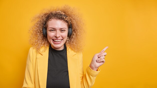 femme aux cheveux bouclés blonds sourit indique agréablement à l'espace de copie vierge écoute de la musique via un casque veste formelle