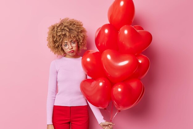 Une femme aux cheveux bouclés bouleversée a une expression triste, un maquillage gâché après avoir pleuré, porte un pantalon rouge décontracté tenant un tas de ballons gonflés en forme de cœur mécontent de célébrer seul un événement festif