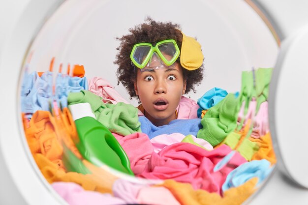 Une femme aux cheveux bouclés choquée occupée à faire la lessive à la maison fait des tâches ménagères quotidiennes dans une machine à laver avec des vêtements sales autour porte des lunettes de plongée en apnée sur le front étant très surprise