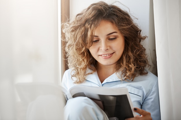 Femme aux cheveux bouclés confortable détendue en vêtements de nuit assis près de la fenêtre et la lecture du magazine de mode