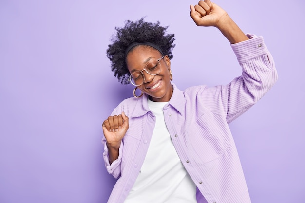 Photo gratuite une femme aux cheveux bouclés danse insouciante lève les mains garde les yeux fermés incline la tête porte des lunettes et une chemise isolée sur un mur violet se déplace sur la piste de danse