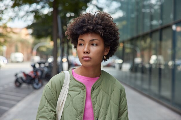 une femme aux cheveux bouclés détourne le regard avec une expression réfléchie pense à quelque chose qui se promène en ville seule vêtue d'une veste profite du beau temps d'automne et d'une belle journée