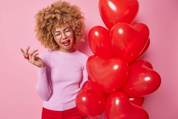 Une femme aux cheveux bouclés frustrée et frustrée pleure de désespoir a l'air lugubre a gâché le maquillage vêtu de vêtements décontractés célèbre la journée des amoureux seule pose près d'un tas de ballons rouges pose à l'intérieur