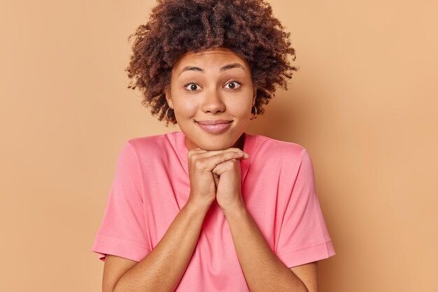 une femme aux cheveux bouclés garde les mains sous le menton écoute des informations avec des expressions curieuses porte un t-shirt rose décontracté isolé sur beige s'intéresse à quelque chose