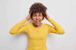 Photo gratuite une femme aux cheveux bouclés gênée attrape la tête s'exclame bruyamment se sent irritée ne sait pas comment résoudre une situation difficile porte un pull jaune isolé sur fond blanc émotions négatives