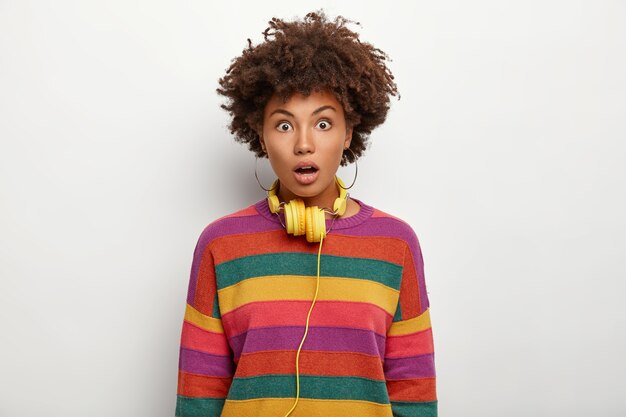 Une femme aux cheveux bouclés horrifiée reçoit des informations terrifiées incroyables, des yeux qui sautent, ouvre la bouche de surprise, porte des écouteurs stéréo autour du cou habillés avec désinvolture isolé sur un mur blanc