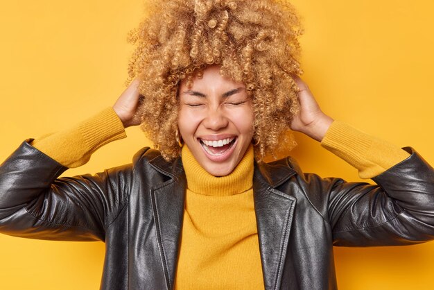 Une femme aux cheveux bouclés, insouciante et positive, rit joyeusement, garde les mains sur la tête, sourit à pleines dents, porte une veste en cuir décontractée isolée sur fond jaune. Concept d'émotions et de sentiments heureux