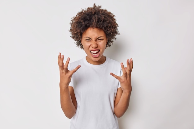 Photo gratuite une femme aux cheveux bouclés irritée lève la main se sent agacée par le conflit s'exclame de la colère exprime des émotions négatives porte un t-shirt décontracté isolé sur fond blanc. comment avez-vous pu faire cela.