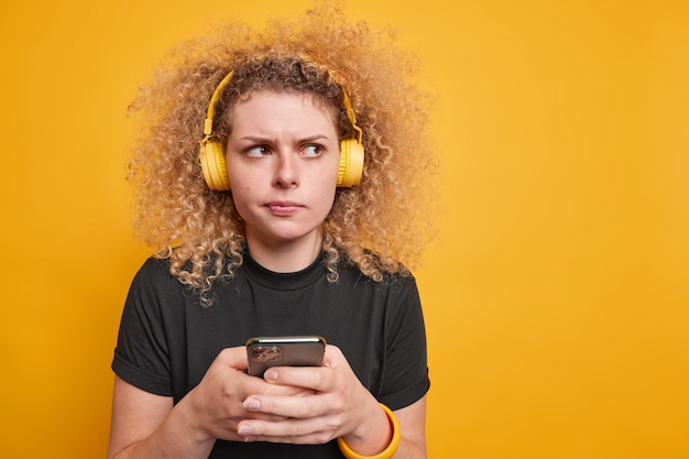 Une femme aux cheveux bouclés mécontente et sérieuse utilise une connexion Internet gratuite pour écouter de la musique en ligne porte des écouteurs sans fil sur les oreilles vêtue d'un t-shirt noir décontracté a une expression réfléchie pose à l'intérieur