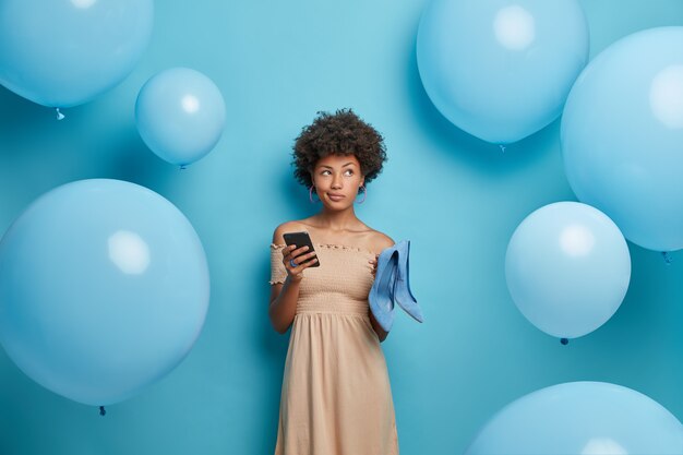 Une femme aux cheveux bouclés à la peau sombre et réfléchie en robe à la mode tient un téléphone portable et envoie des invitations à des amis.La soirée à thème ballons choisit les meilleures chaussures à porter entourées de ballons gonflés