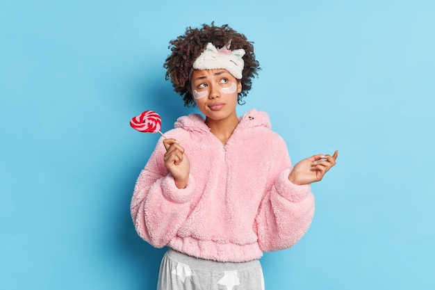 Une femme aux cheveux bouclés et perplexe réfléchie soulève la paume concentrée quelque part porte pensivement un masque de sommeil et un pyjama tient des bonbons sucrés sur un bâton applique des produits de nuit de soins de la peau sur le visage