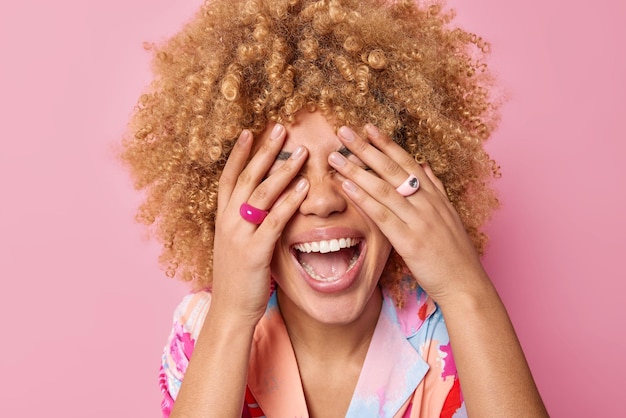 Une femme aux cheveux bouclés positive couvre les yeux avec les mains attend des rires surprises joyeusement a une humeur optimiste garde la bouche ouverte montre des dents blanches isolées sur fond rose Les gens et les émotions sincères