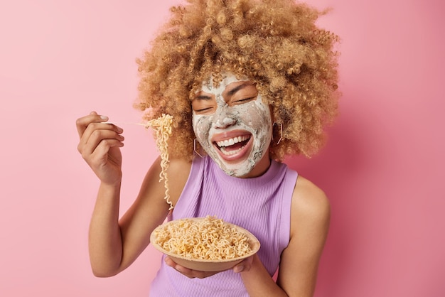 Une femme aux cheveux bouclés ravie mange des pâtes avec appétit rit joyeusement applique un masque facial de beauté pour le traitement de la peau vêtue d'un t-shirt violet isolé sur fond rose