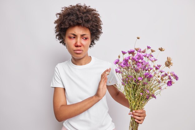 une femme aux cheveux bouclés refuse d'avoir un bouquet de fleurs sauvages allergique au pollen a l'air malheureuse a les yeux rouges et le nez habillé avec désinvolture pose sur un mur blanc