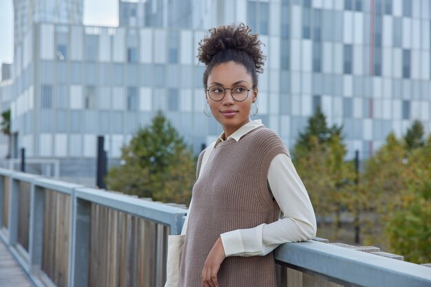 femme aux cheveux bouclés vêtue de vêtements à la mode lunettes rondes regarde loin considère quelque chose