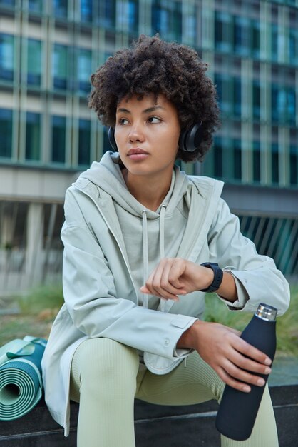 une femme aux cheveux bouclés vêtue de vêtements de sport vérifie l'heure sur la smartwatch a soif tient une bouteille d'eau écoute la piste audio via un casque pose contre un bâtiment urbain