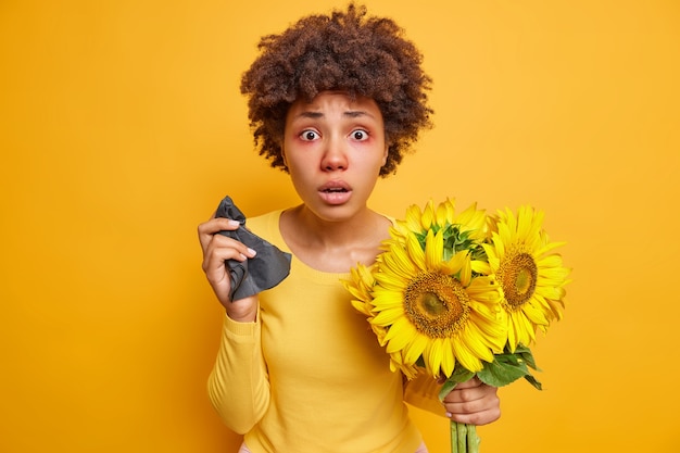 femme aux cheveux bouclés yeux gonflés rouges tient des éternuements de serviette en raison de l'allergie aux tournesols étant malade pose à l'intérieur sur jaune vif