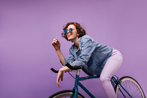 Femme aux cheveux courts insouciante assise sur un vélo. Heureuse fille caucasienne avec une coiffure ondulée exprimant des émotions positives.