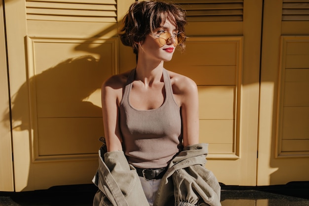 Femme aux cheveux courts en lunettes de soleil posant sur les portes jaunes. Femme bouclée en maillot de corps avec veste regarde ailleurs sur les portes jaunes