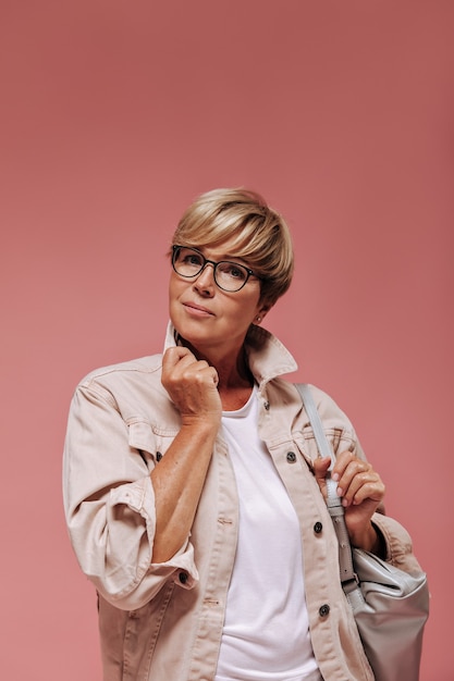 Femme aux cheveux courts modernes, boucles d'oreilles et lunettes cool en veste à la mode et t-shirt léger posant avec sac gris sur fond rose.