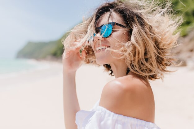 Femme aux cheveux courts romantique avec beau sourire posant sur la nature floue. Charmante femme bronzée à lunettes de soleil en riant tout en se reposant sur une plage exotique.