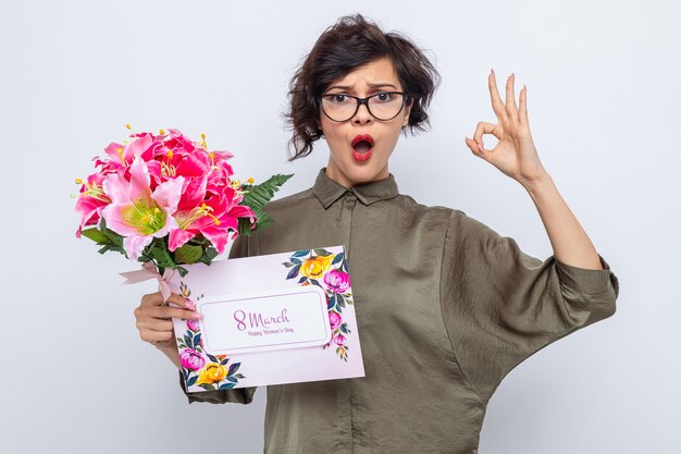 Femme aux cheveux courts tenant une carte de voeux et un bouquet de fleurs regardant la caméra confuse et surprise en train de faire signe ok célébrant la journée internationale de la femme le 8 mars debout sur fond blanc