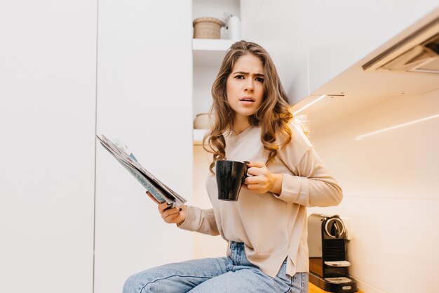 Femme aux cheveux longs émotionnelle porte chemise beige boire du café dans sa cuisine