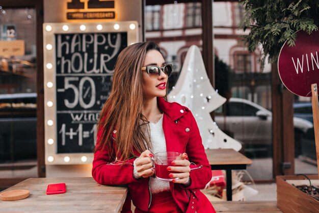 Femme aux cheveux longs en lunettes de soleil noires debout dans la rue avec une tasse de thé