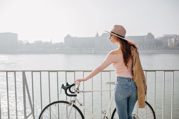 Femme aux cheveux longs réfléchie au chapeau debout près de vélo et profiter de la vue sur la rivière en journée ensoleillée