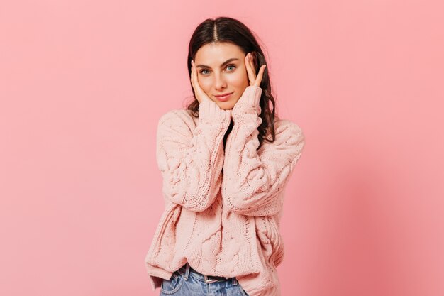 Femme aux cheveux noirs aux yeux bleus touche doucement son visage. Fille en pull surdimensionné et jeans regardant la caméra sur fond rose.