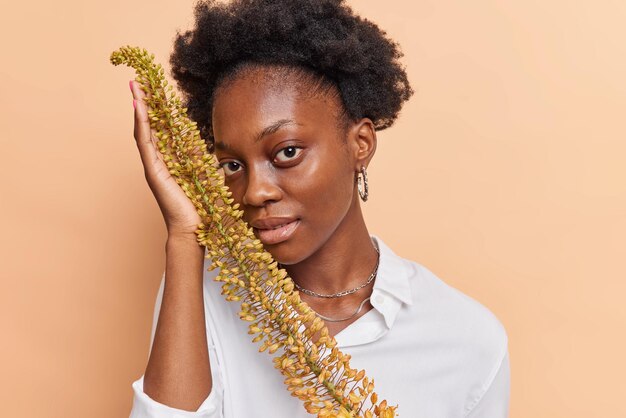 une femme aux cheveux noirs bouclés tient une plante exotique près du visage regarde directement la caméra porte une chemise blanche a un teint bien soigné isolé sur beige